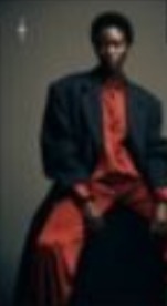 Figure in a dark coat and red trousers seated against grey